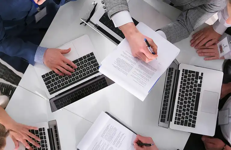 Overhead view of professionals working on laptops and reviewing printed documents at a white table.