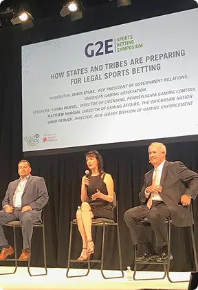 Three panelists seated on stage under a large screen displaying "G2E Sports Betting Symposium" and discussion topic about states and tribes preparing for legal sports betting.