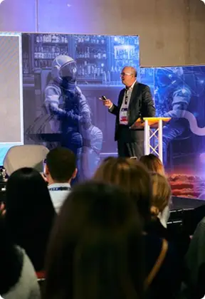 A man in a suit presenting on stage with astronaut images on large screens behind him.