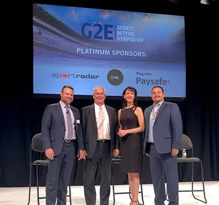 Four professionally dressed individuals standing on stage under a screen displaying "G2E Sports Betting Symposium" and sponsor logos.