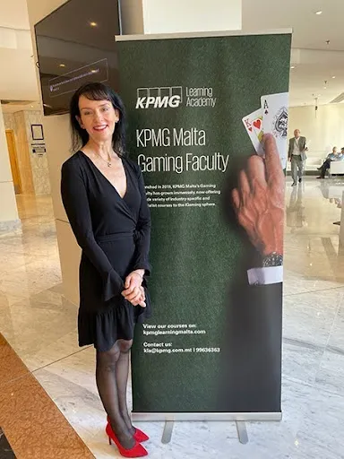 Professional woman in black dress and red heels standing beside KPMG Malta Gaming Faculty banner.