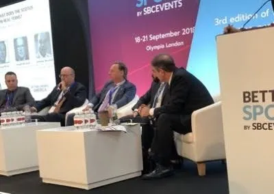 Panel of four men in business attire seated on stage at a conference with a purple backdrop.