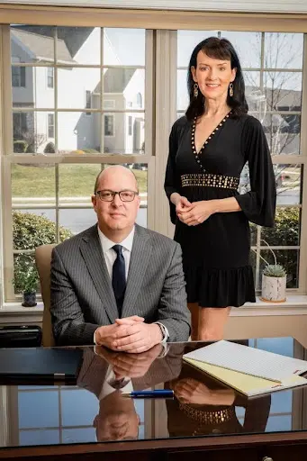 Two professionally dressed individuals, a seated man in a suit and a standing woman in a black dress, in a bright office with large windows.