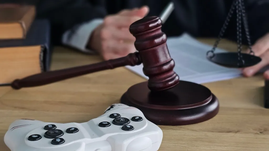 Wooden judge's gavel and white video game controller on desk with legal books and scales.