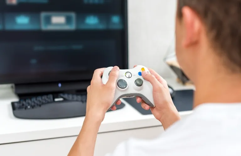 Person holding a white video game controller in front of a monitor and keyboard.