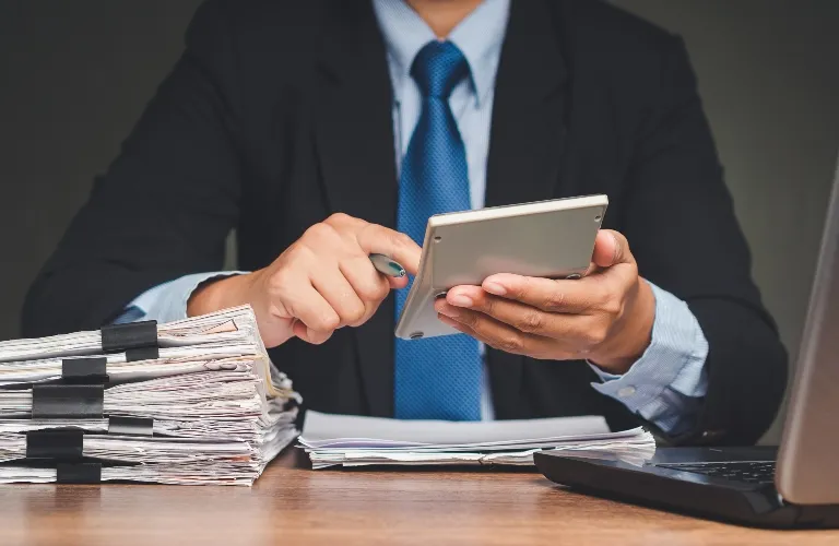 Person in business attire using calculator with stacks of documents and laptop on desk.